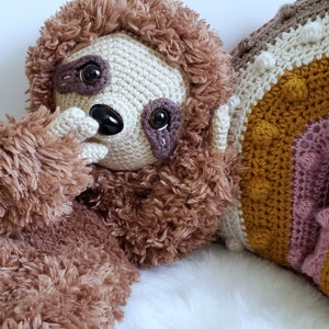 May include: A brown and beige crocheted sloth plush toy with a white background. The sloth is holding its paw to its face and has brown eyes. The sloth is sitting next to a crocheted rainbow pillow with brown, beige, and yellow stripes.