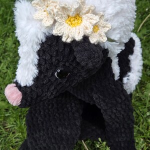 May include: A handmade plush skunk toy with a white fluffy head and a black body. The skunk wears a daisy crown. The toy is made of soft, textured material and has a pink nose. The skunk is sitting on green grass.