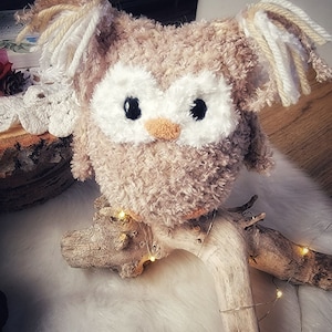 May include: A brown and white crocheted owl with large eyes and a fluffy texture. The owl is sitting on a piece of driftwood with white lights wrapped around it.