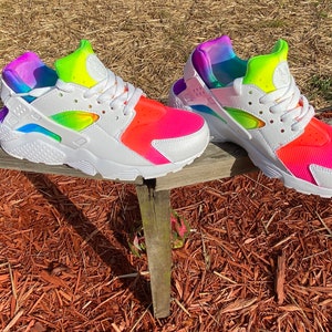 May include: A pair of white Nike Air Huarache sneakers with a rainbow tie-dye design. The sneakers have a white midsole and a black outsole.