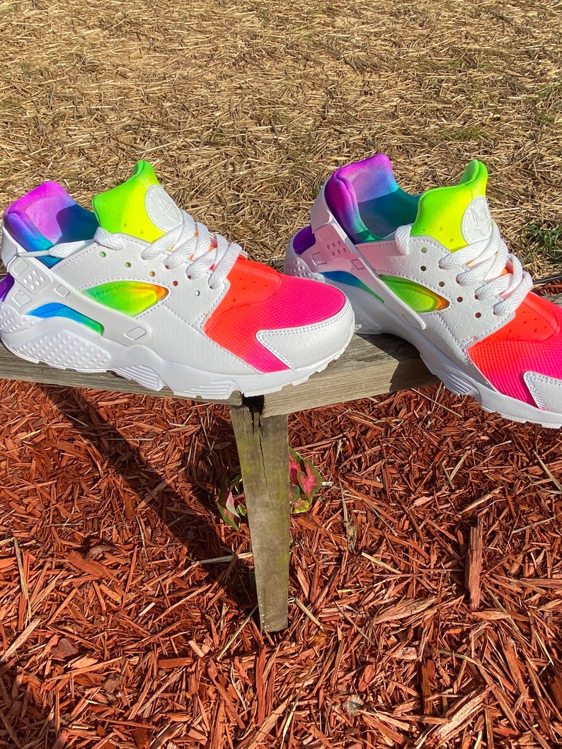 May include: A pair of white Nike Air Huarache sneakers with a rainbow tie-dye design. The sneakers have a white midsole and a black outsole.