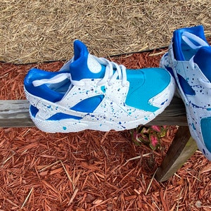 May include: A pair of white sneakers with blue and turquoise accents. The sneakers have a splatter paint design and are sitting on a bed of wood chips.