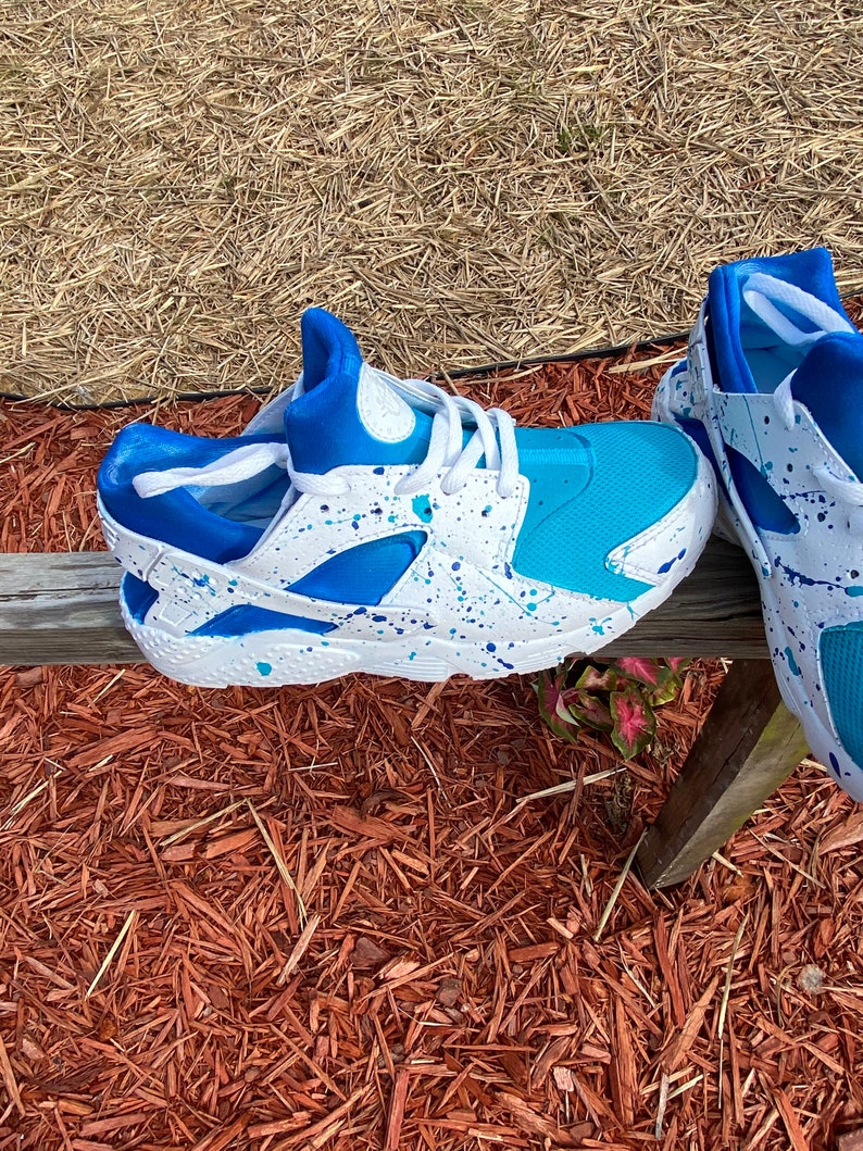 May include: A pair of white sneakers with blue and turquoise accents. The sneakers have a splatter paint design and are sitting on a bed of wood chips.