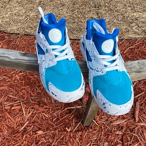 May include: A pair of white sneakers with blue and white paint splatter. The sneakers have a blue mesh upper and a white sole.