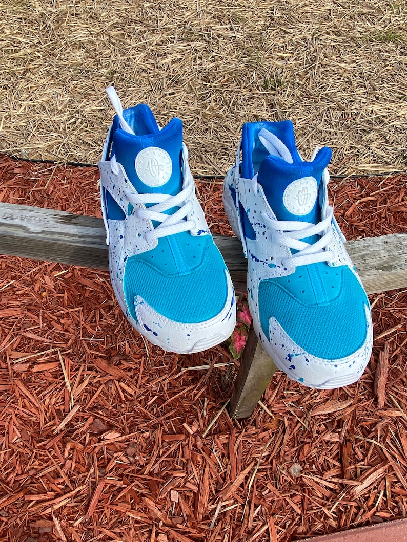 May include: A pair of white sneakers with blue and white paint splatter. The sneakers have a blue mesh upper and a white sole.