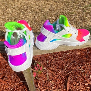 May include: A pair of white Nike Huarache sneakers with a rainbow tie-dye design. The sneakers have a white swoosh logo on the side and a white Nike logo on the back.