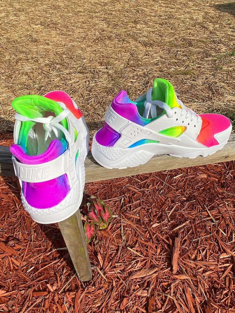 May include: A pair of white Nike Huarache sneakers with a rainbow tie-dye design. The sneakers have a white swoosh logo on the side and a white Nike logo on the back.