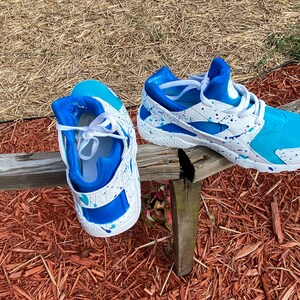 May include: A pair of white sneakers with blue and turquoise accents. The sneakers have a white base with blue and turquoise paint splatters. The sneakers are sitting on a wooden surface.