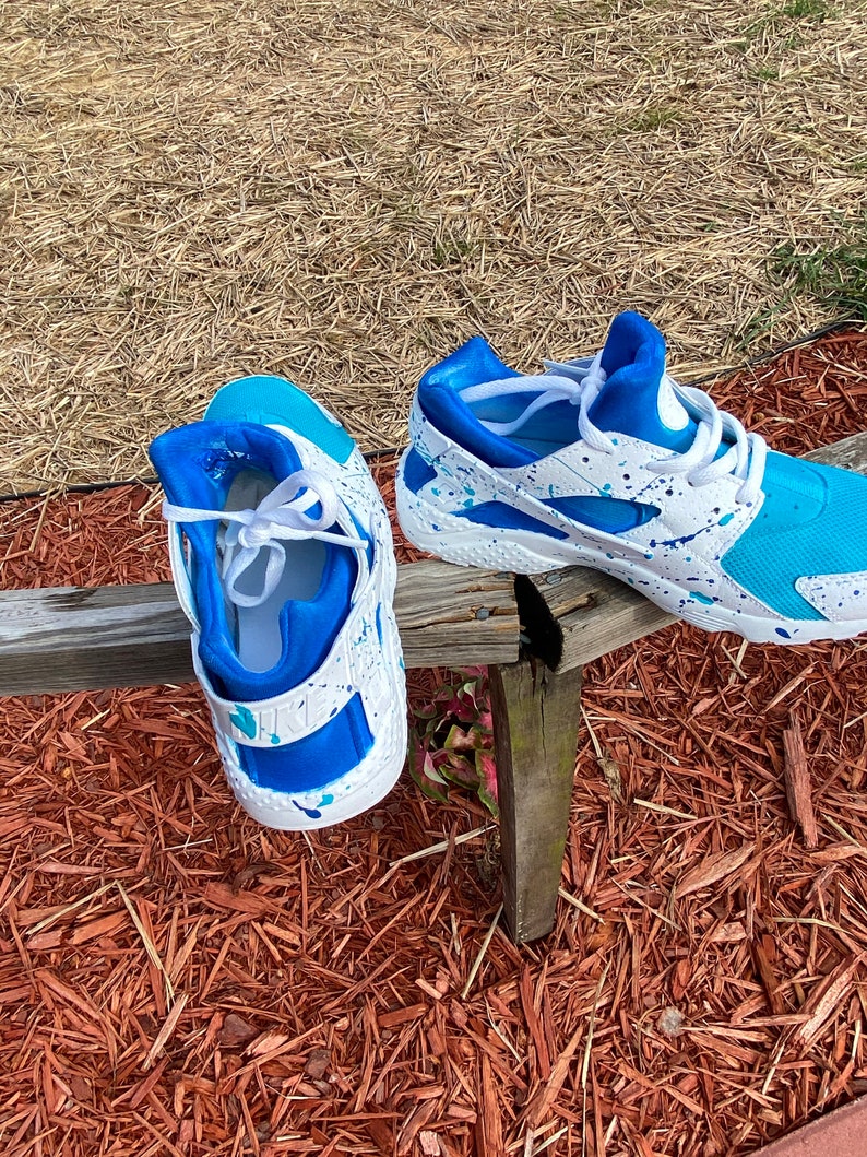 May include: A pair of white sneakers with blue and turquoise accents. The sneakers have a white base with blue and turquoise paint splatters. The sneakers are sitting on a wooden surface.