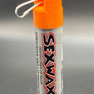 Peut inclure: Tube de baume à lèvres orange et argent avec un mousqueton argenté. Le tube est étiqueté "SEXWAX DERMATONE® LIP BALM SUNSCREEN AND LIP PROTECTOR".
