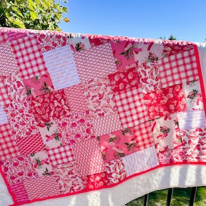 May include: A patchwork quilt with a variety of pink patterns, including gingham, butterflies, and floral prints. The quilt has a white backing and a bright pink border. The quilt is displayed outdoors.