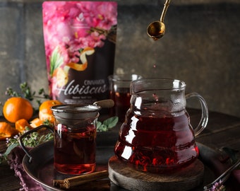 May include: A glass carafe and a glass mug with a metal strainer containing deep red hibiscus tea. A gold spoon drips liquid into the carafe. Nearby are cinnamon sticks, fresh orange segments, and a pouch labelled Cinnamon Hibiscus.