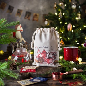 May include: A white canvas bag with a Christmas-themed design featuring a teapot, candy canes, and the text "Merry Christmas". The bag is sitting on a wooden table with a red mug, tea bags, and a small bowl of cranberries.