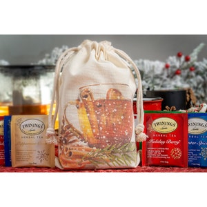 May include: A white canvas drawstring bag with a festive design of a mug of hot beverage with cinnamon sticks and snowflakes. The bag contains three boxes of Twinings herbal tea: Peppermint Cocoa, Holiday Berry, and Winter Spice.