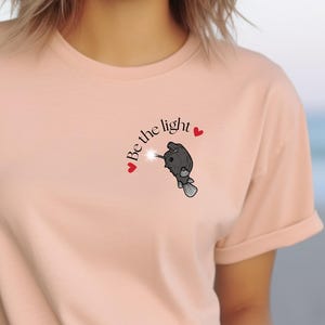 以下が含まれることがあります： 薄いピンクのTシャツに、杖を持った鳥の白黒のイラストと「Be the light」という文字、そして赤いハートが2つ描かれています。