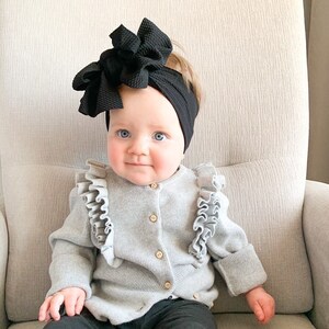 May include: A baby girl wearing a gray cardigan with ruffles, black pants, and a black bow headband. The baby is sitting in a gray chair.