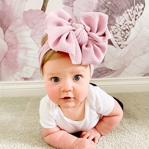 May include: A baby wearing a pink knotted headband with a large bow. The baby is lying on its stomach on a light beige carpet.