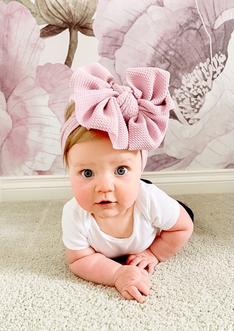 May include: A baby wearing a pink knotted headband with a large bow. The baby is lying on its stomach on a light beige carpet.