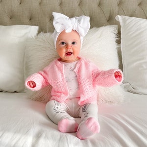 May include: A baby wearing a pink knitted cardigan and white leggings with a white bow headband. The baby is sitting on a white bed.