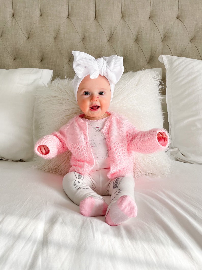 May include: A baby wearing a pink knitted cardigan and white leggings with a white bow headband. The baby is sitting on a white bed.
