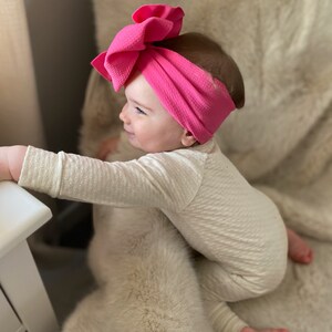 May include: A baby wearing a white long-sleeved onesie and a bright pink knotted headband with a large bow. The baby is sitting on a fluffy white rug.