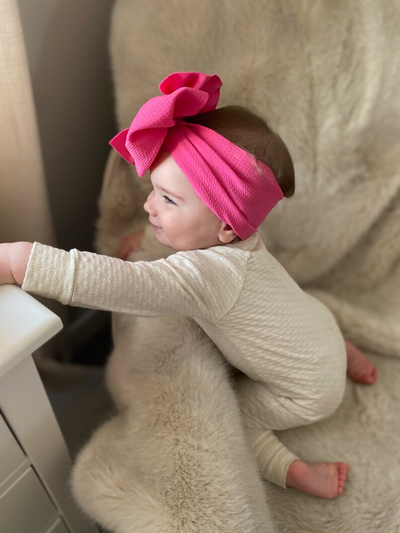 May include: A baby wearing a white long-sleeved onesie and a bright pink knotted headband with a large bow. The baby is sitting on a fluffy white rug.