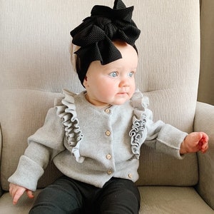 May include: A baby wearing a gray cardigan with ruffles, black leggings, and a large black bow headband. The baby is sitting in a chair and looking to the right.