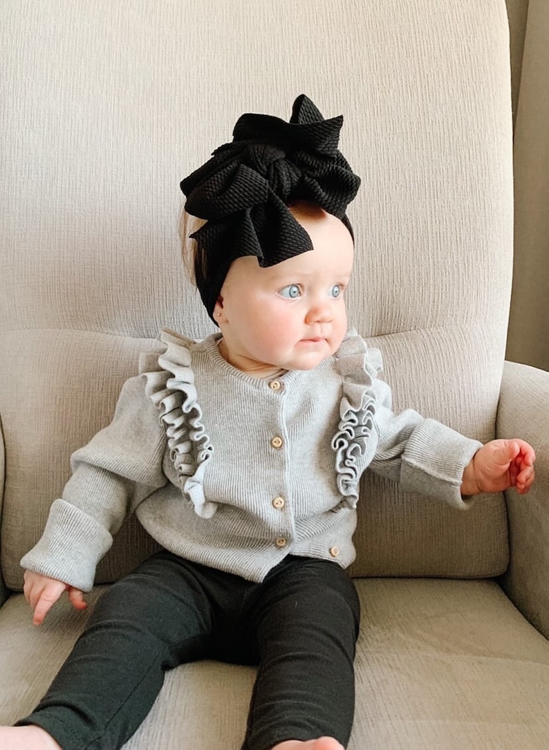 May include: A baby wearing a gray cardigan with ruffles, black leggings, and a large black bow headband. The baby is sitting in a chair and looking to the right.