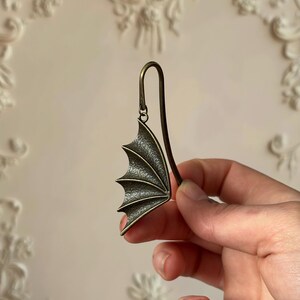 May include: A bronze-colored bookmark shaped like a dragon wing. The wing has a textured surface with detailed ridges and a curved metal stem. The bookmark is held against a light-colored background.