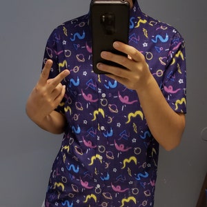 May include: A short-sleeved button-down shirt in a dark blue color, patterned with colorful cartoon-like shapes. The design includes pink, yellow, and blue worm-like figures, planets, stars, and geometric shapes. The shirt has a classic collar and a button-up front.