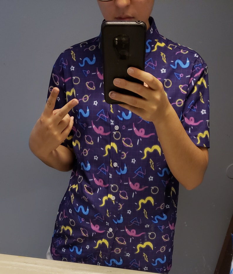 May include: A short-sleeved button-down shirt in a dark blue color, patterned with colorful cartoon-like shapes. The design includes pink, yellow, and blue worm-like figures, planets, stars, and geometric shapes. The shirt has a classic collar and a button-up front.