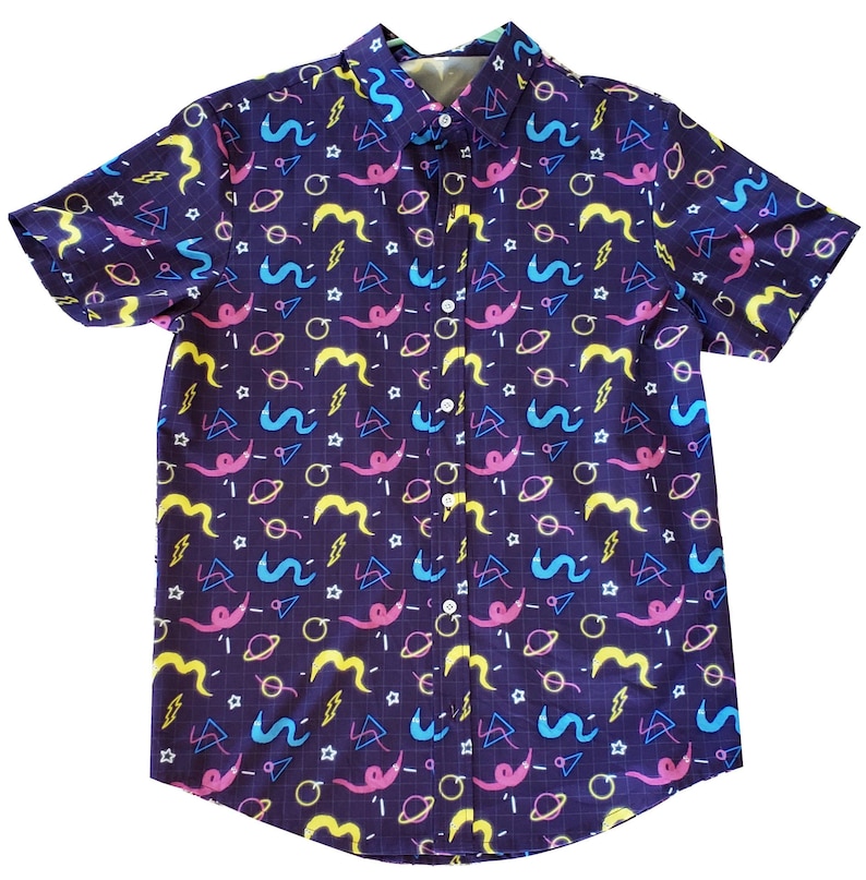 May include: Short-sleeved button-down shirt with a dark purple background and a colorful pattern. The pattern includes yellow, pink, blue, and white shapes, including squiggles, planets, and lightning bolts. The shirt has a classic collar and white buttons.