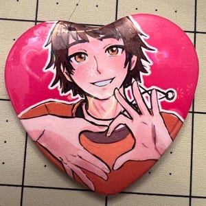 May include: Heart-shaped button featuring an anime-style illustration of a person making a heart shape with their hands. The background is pink with small star-like designs. The person has brown hair and is wearing an orange shirt.