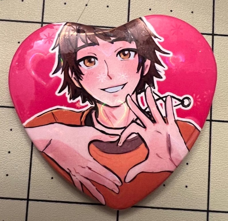 May include: Heart-shaped button featuring an anime-style illustration of a person making a heart shape with their hands. The background is pink with small star-like designs. The person has brown hair and is wearing an orange shirt.