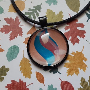 Puede incluir: Un collar negro con un colgante redondo de vidrio que presenta un diseño de remolino azul y rosa. El colgante está engastado en un marco de metal negro. El collar está fotografiado sobre un fondo de hojas de otoño impresas.