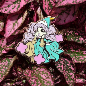 May include: A metal enamel pin featuring an anime-style character with voluminous light purple hair, wearing a teal pointed hat and a matching teal cape over a tan outfit. The character has hands on cheeks and is surrounded by three pink cloud shapes.
