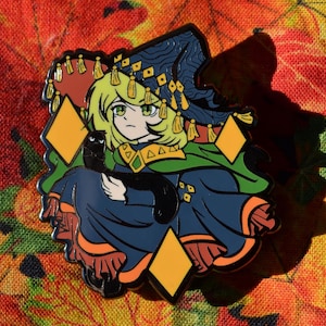 May include: A metal enamel pin featuring an anime-style character with blonde hair wearing a dark blue and green robe with a matching pointed hat. The character holds a small black cat, surrounded by three orange diamond shapes with black outlines.