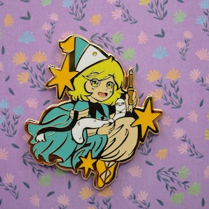 May include: A gold enamel pin depicting a cartoon witch with yellow hair, wearing a teal dress and a pointed hat. She holds a white cat and a pen, riding a broom with star accents. The pin is set against a purple floral background.