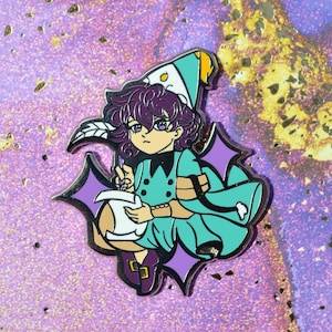 May include: A metal enamel pin featuring a character with dark purple hair wearing a teal outfit and a matching pointed hat. The character holds a white quill and paper, surrounded by four-pointed purple star shapes with black outlines.