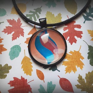 May include: A round pendant necklace featuring a black metal frame and a glass cabochon. The center design displays a swirling abstract pattern with vibrant blue and deep red colors against a muted tan background, suspended on a black cord.