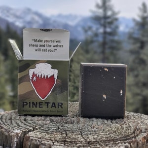 May include: A bar of dark brown pine tar soap with a white label that reads "PINETAR Natural Hand-Crafted Small Batch". The soap is sitting on a wooden surface with a mountain background. The label also includes the quote "Make yourselves sheep and the wolves will eat you!"