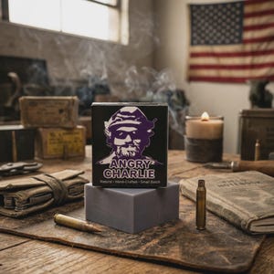 May include: A black box with the words "ANGRY CHARLIE" and a purple illustration of a man. The box sits on a gray rectangular soap bar. The words "Natural - Hand-Crafted - Small Batch" are below the name. The background includes a lit candle and an American flag.