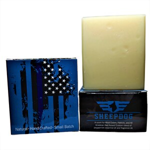 May include: A bar of soap in a white box with a black and blue American flag design. The box has the text "Natural Hand-Crafted Small Batch" on the front. The soap is in a clear plastic wrapper. The box also has the text "SHEEPDOG" and "A scent for Meat Eaters, Patriots, and All American Bad Asses! Custom blend of peppermint essential oil and fragrance oil."
