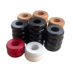 May include: A collection of wooden game pieces in various colors. There are stacks of black, red, and white pieces, along with natural wood-toned pieces. The pieces are circular with a hole in the center, likely for a ring toss or similar game.