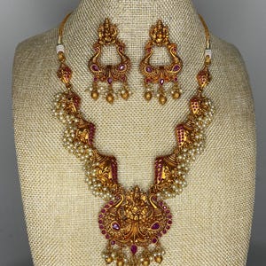 May include: A gold necklace and earrings set featuring a peacock design with pink and red stones. The necklace has a large pendant with a peacock design and a string of pearls.