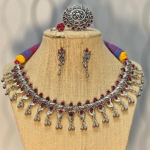 May include: A silver-toned jewelry set featuring a necklace, earrings, and two rings, all adorned with red gemstones. The necklace has a detailed, ornate design with dangling accents. The rings include a large statement ring and a smaller ring. The jewelry is displayed on a mannequin.