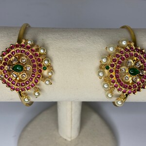 May include: A pair of gold bangles with intricate designs featuring pink and white stones, green accents, and pearl embellishments.