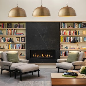 May include: A living room with a fireplace, two armchairs, and a coffee table. The fireplace has a black surround and a gas fire. There are three brass pendant lights hanging from the ceiling. The room is decorated with bookshelves filled with books.
