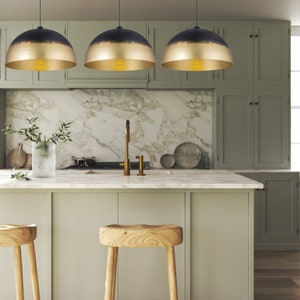 May include: Three gold and black dome-shaped pendant lights hang over a kitchen island with a marble countertop and green cabinets. Two wooden stools are positioned in front of the island.