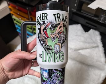 May include: White stainless steel tumbler with a black lid and handle. The tumbler features a green, black, and white graphic of characters from the movie "The Nightmare Before Christmas" with the text "Never Trust The Living". The bottom of the tumbler has a repeating pattern of the same text and images.